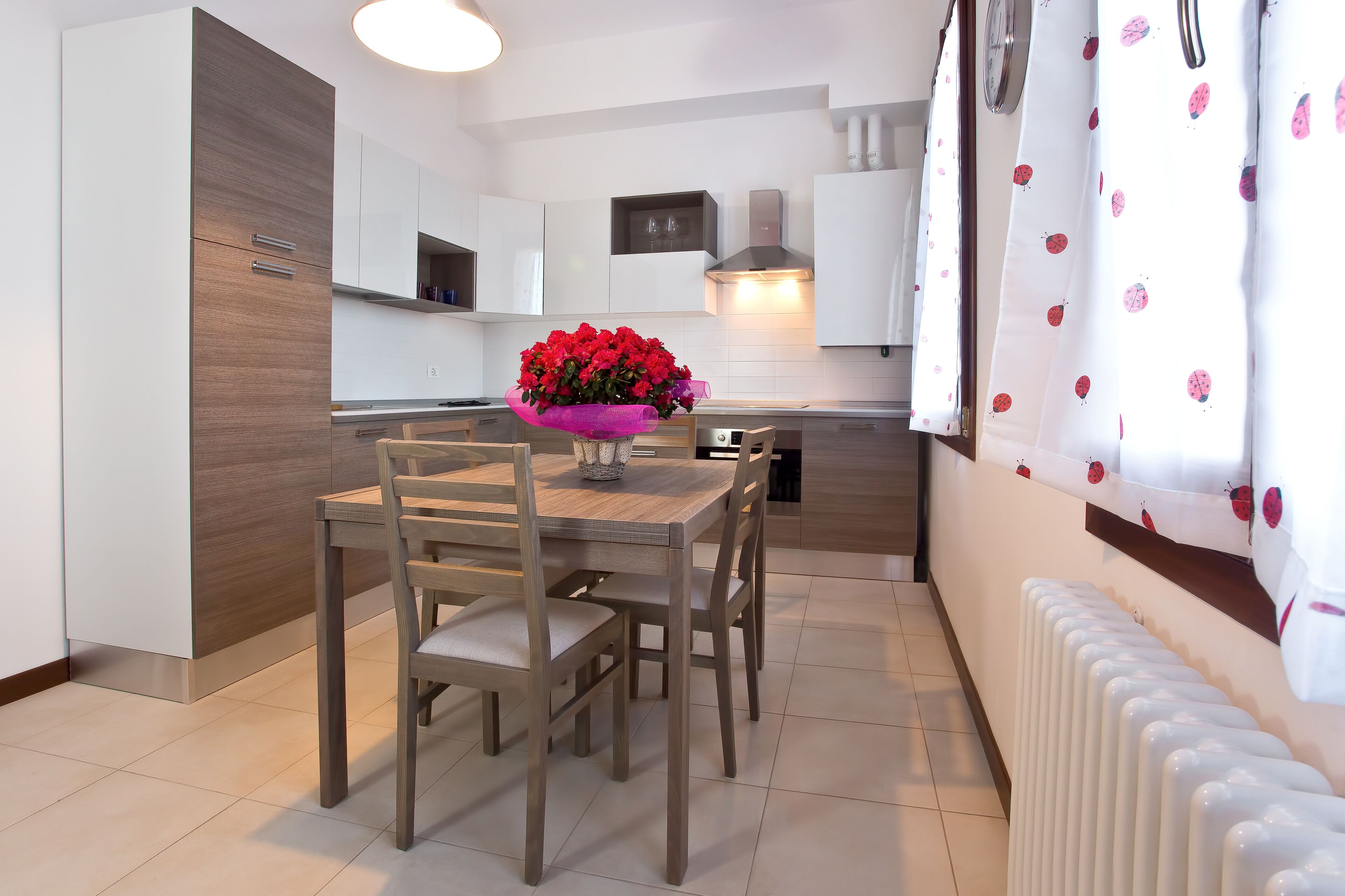 A modern kitchen with white cabinets, a wooden dining table, and a vase of red flowers.