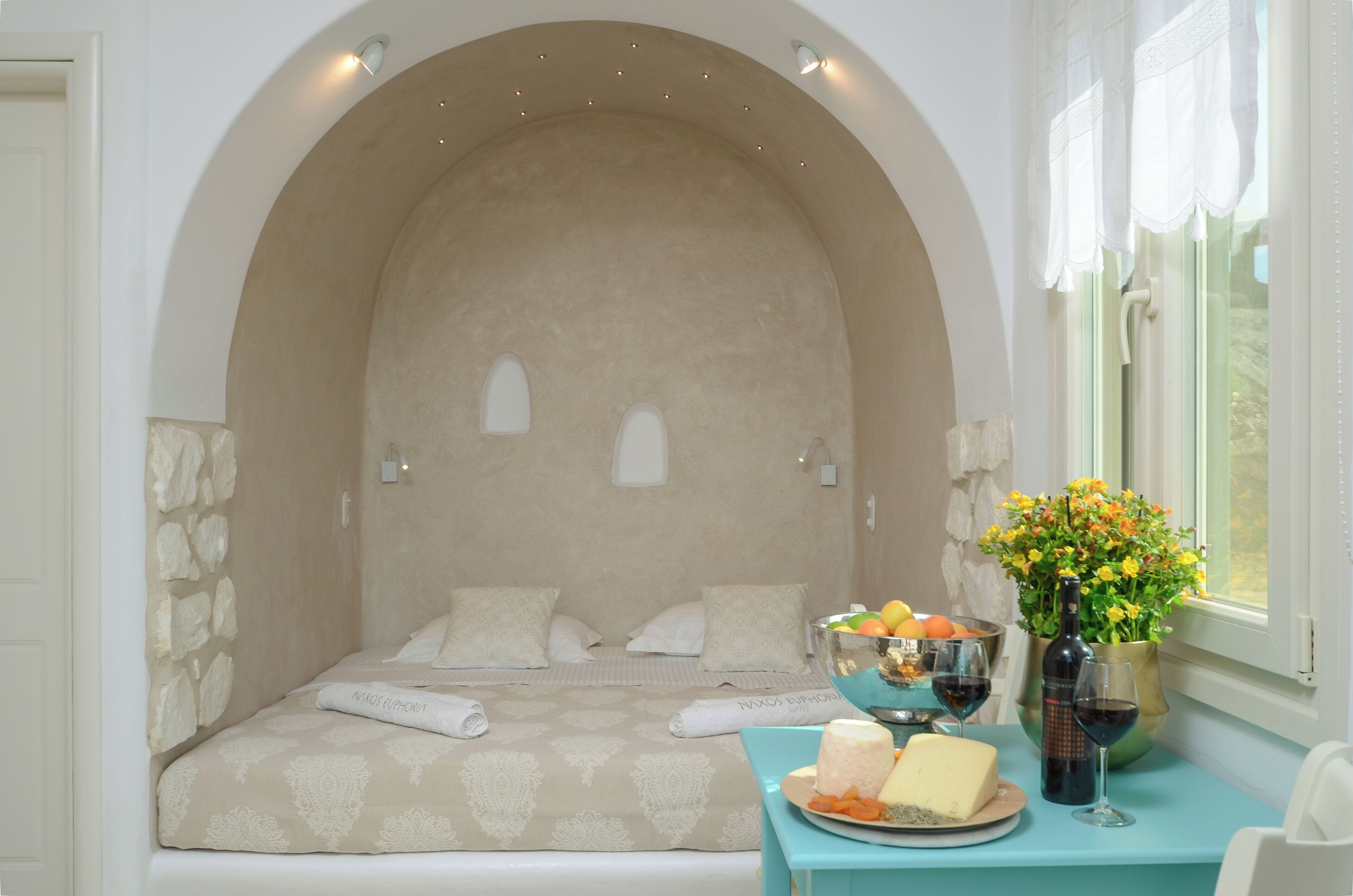 A bedroom with a stone wall, a bed with a patterned bedspread, a table with a fruit bowl, wine glasses, and a bottle.