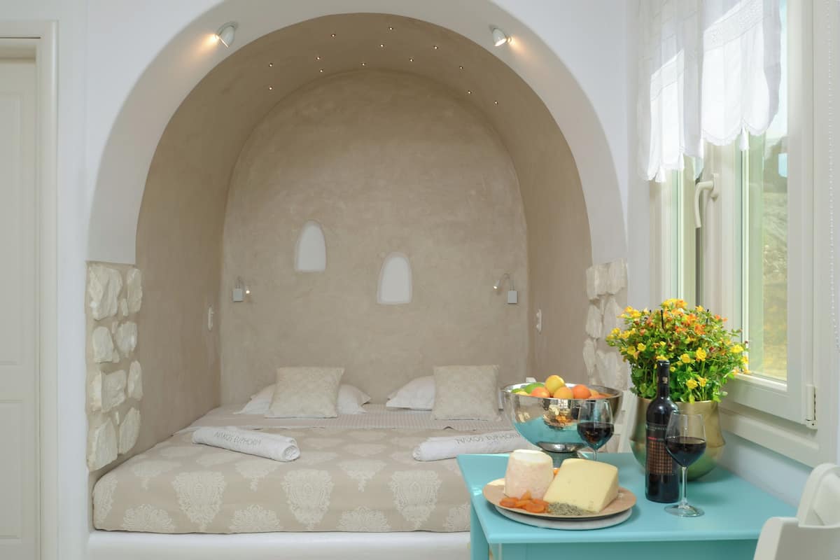 Un dormitorio con pared de piedra, una cama con colcha estampada, una mesa con frutero, copas de vino y una botella.