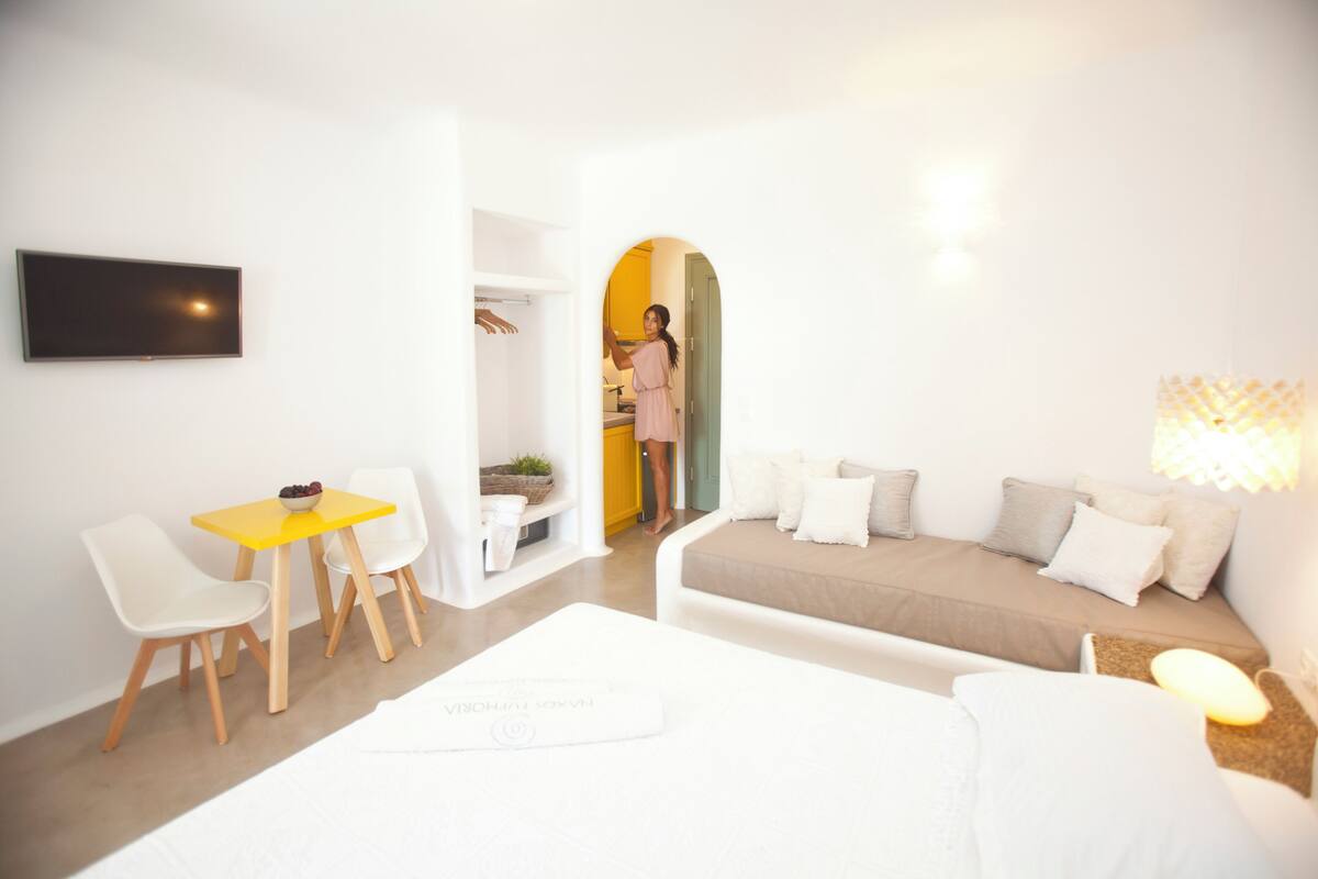 A modern hotel room with a bed, a sofa, a dining table with chairs, and a TV mounted on the wall.