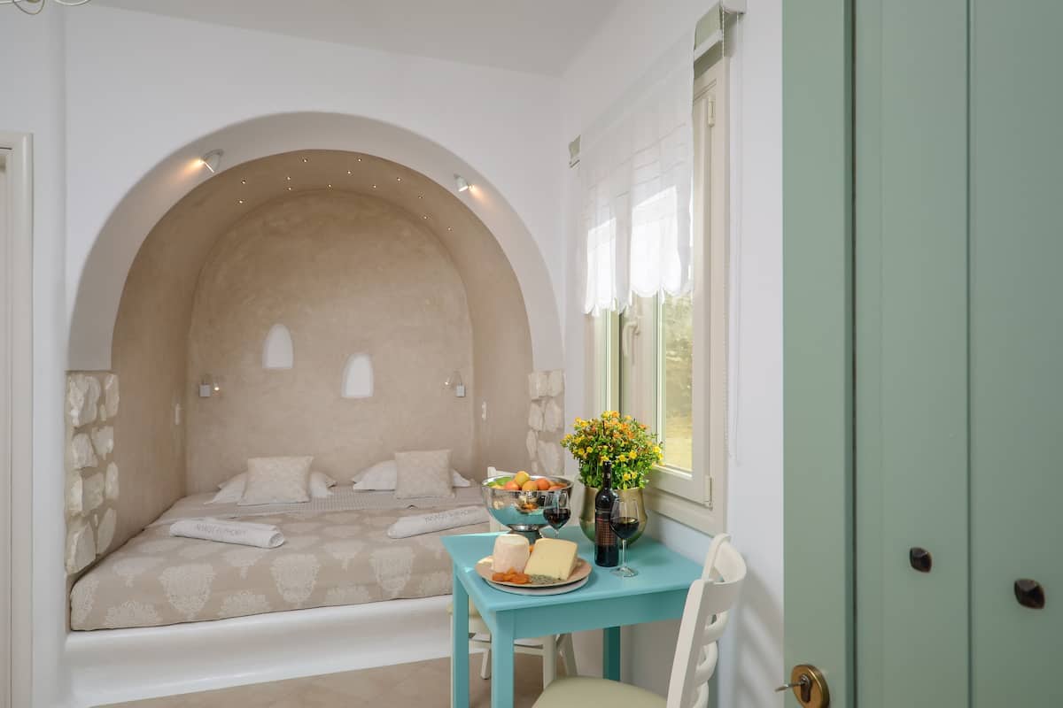 Un dormitorio acogedor con pared de piedra, cama con colcha estampada, una mesita con frutero, queso y copas de vino, y un jarrón con flores.