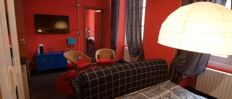 A hotel room with a red wall, a black and white checkered sofa, a blue dresser, a television mounted on the wall, and a window with curtains.