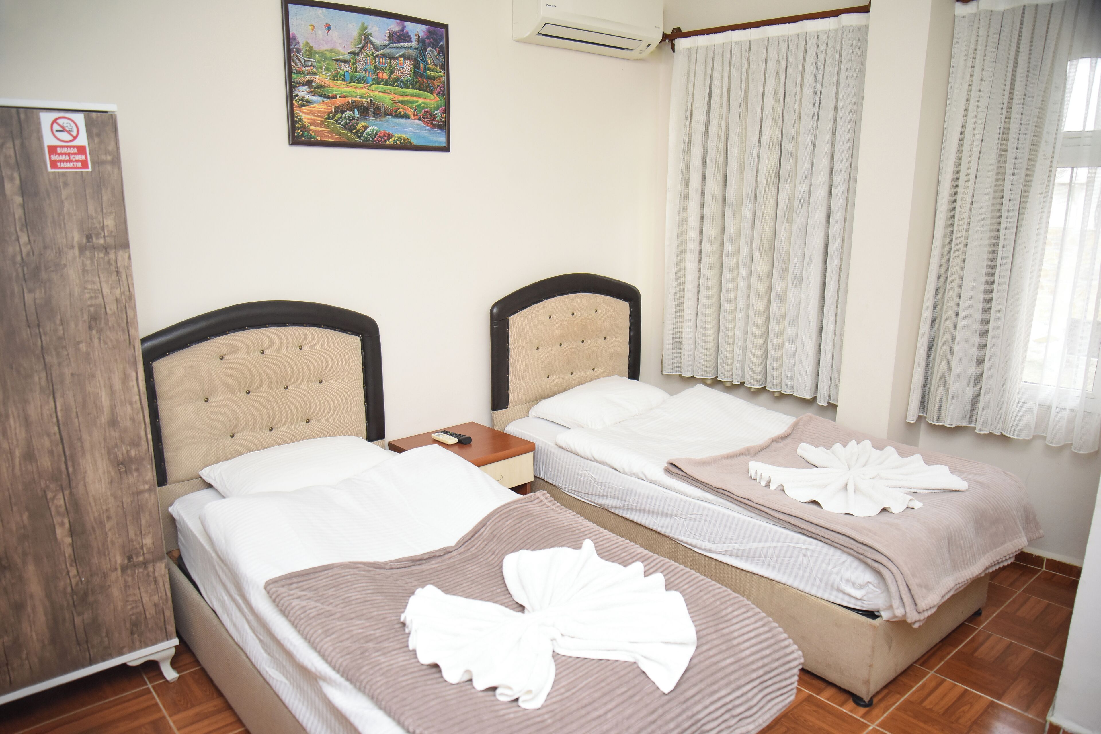 A hotel room with two beds, a wooden wardrobe, a painting on the wall, and a window with curtains.