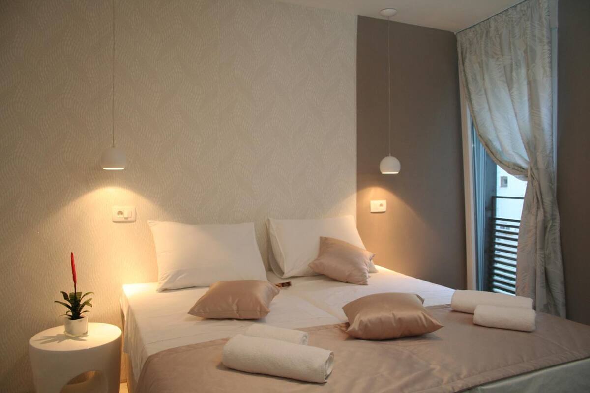 A neatly made bed with pillows and rolled towels, a small white side table with a plant, and a window with curtains.