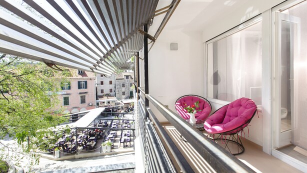 A balcony with two pink chairs, a view of a street below, and a building with a tiled roof. - Guest House Teatrin Split (Split)