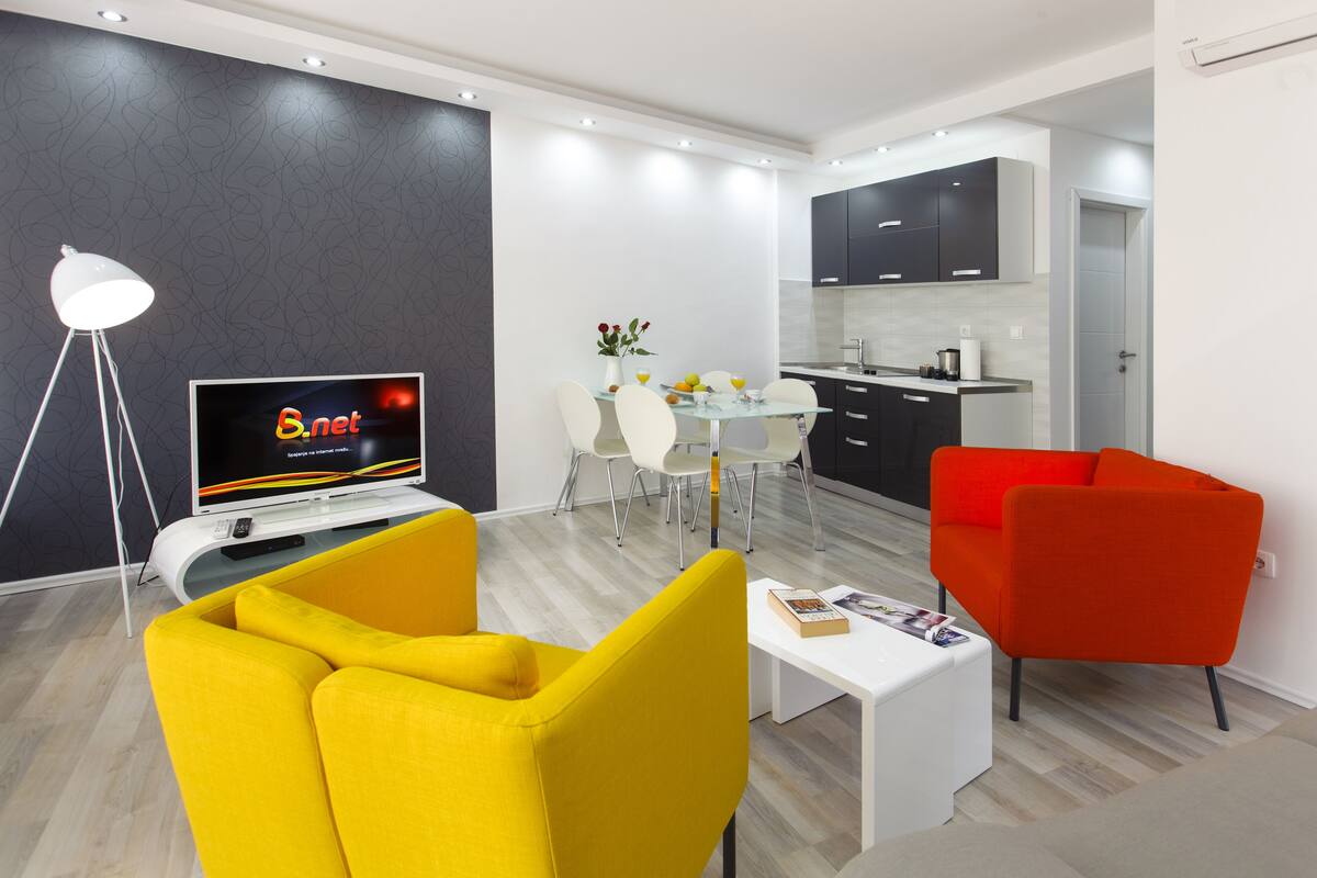 A modern living room with a TV, yellow and red chairs, a dining table with chairs, and a kitchen area.