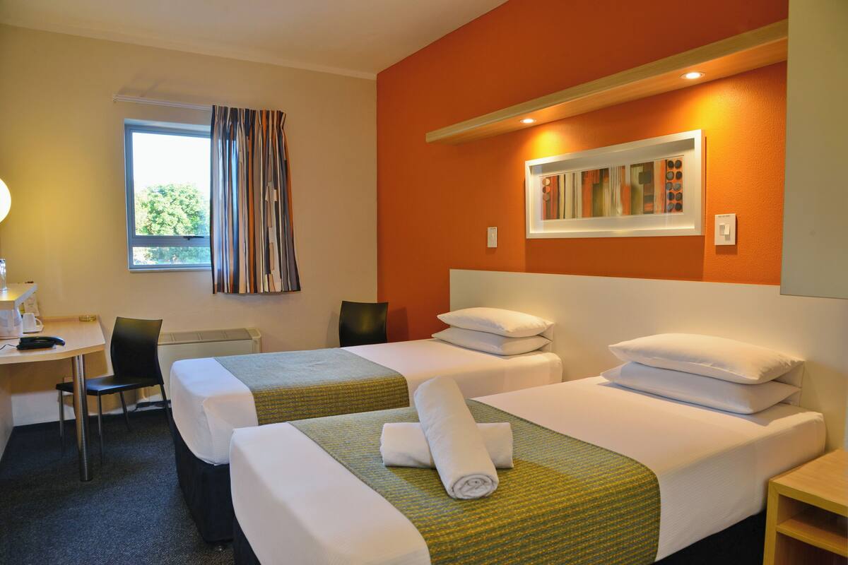 Habitación de hotel con dos camas, un escritorio, una silla y un ventanal con cortinas.