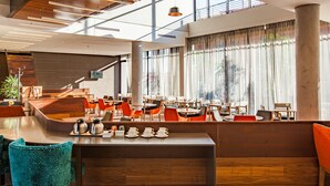 A modern hotel lobby with a wooden reception desk, seating area with orange chairs, and large windows with sheer curtains.