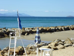 A beach with rocky terrain, a white lounge chair, a blue umbrella, and a white table. - Baia Etrusca (Piombino)