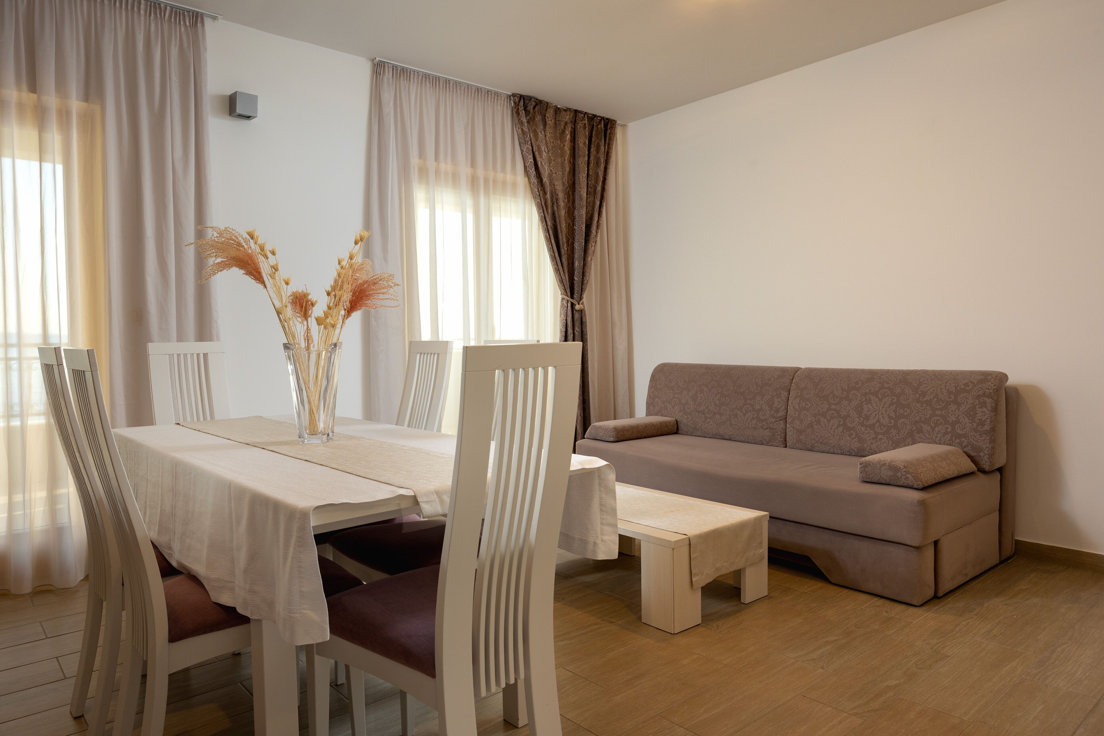 Ein modernes Wohnzimmer mit Esstisch, weißen Stühlen, einer Couch und einer Vase mit getrockneten Blumen.