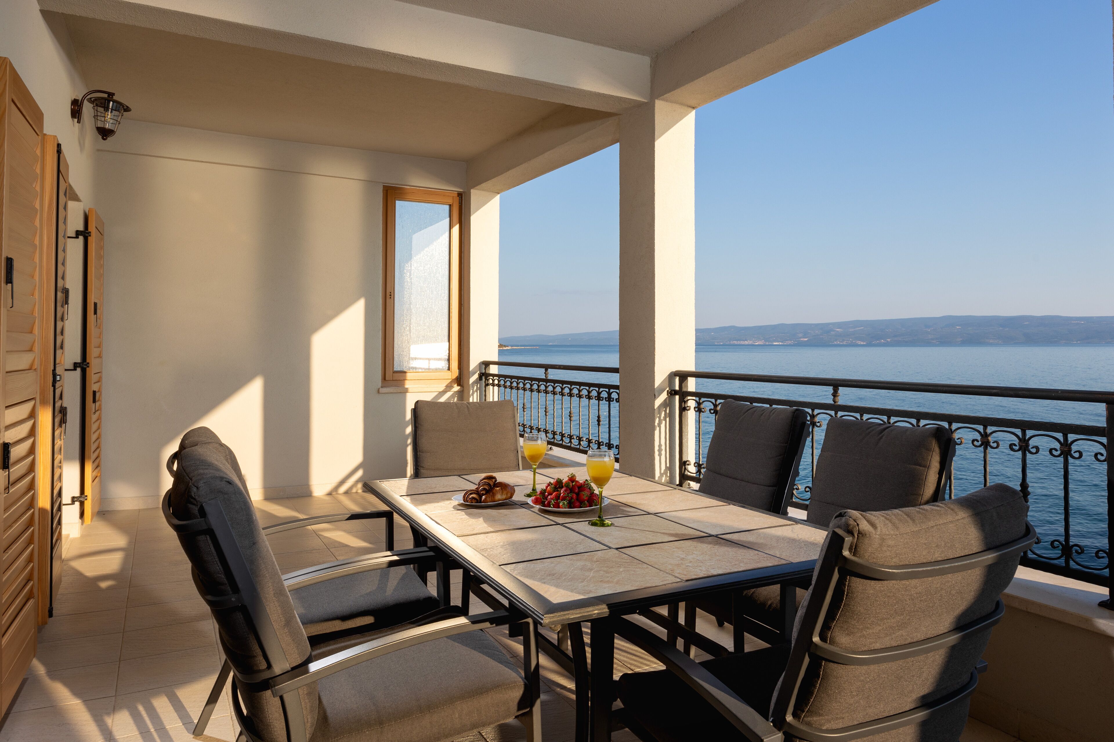 A balcony with a table set for two, featuring a plate of croissants and a glass of orange juice, overlooking a body of water.