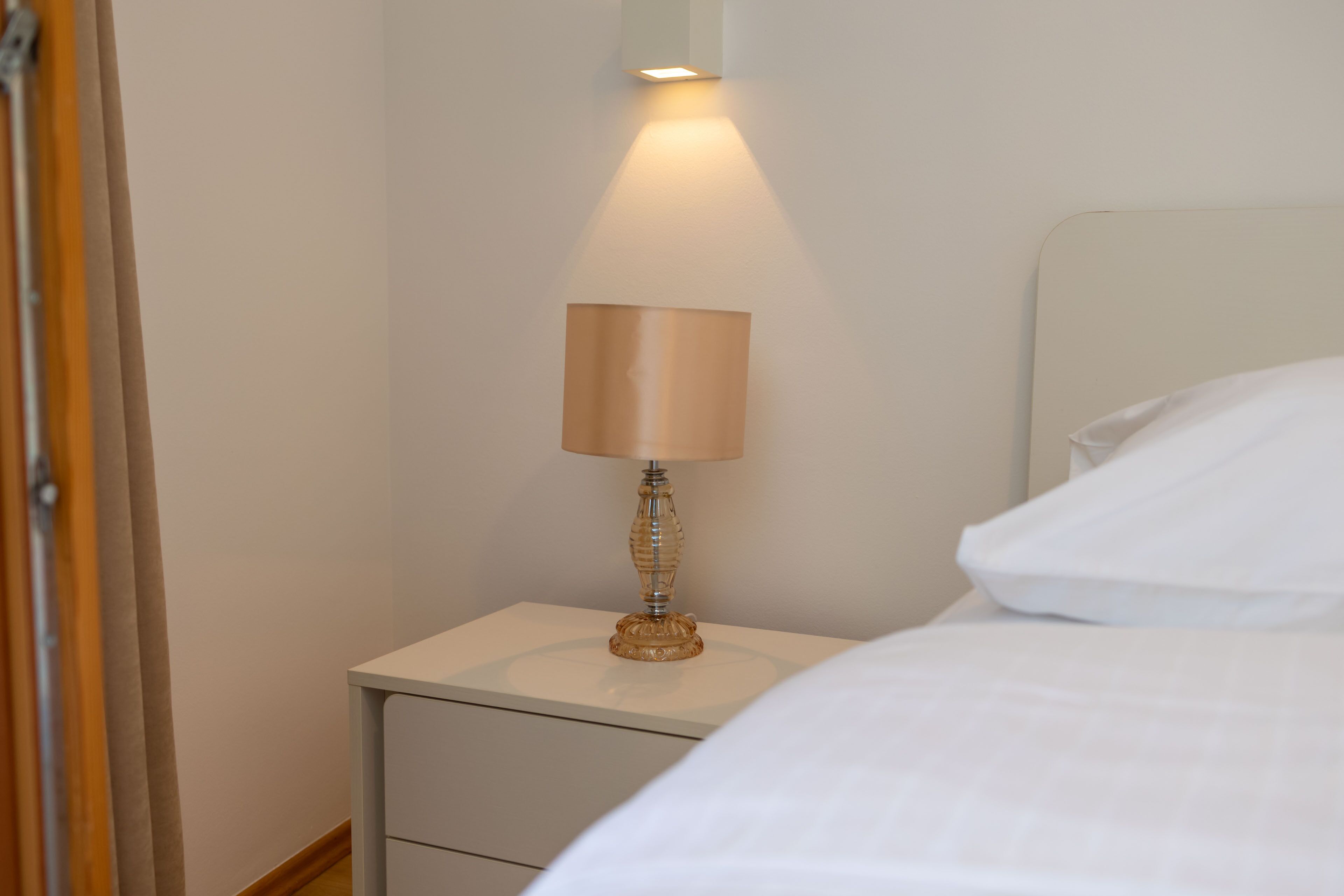 Una cama bien tendida con sábanas blancas, una lámpara de noche y una mesita de noche blanca con cajones.