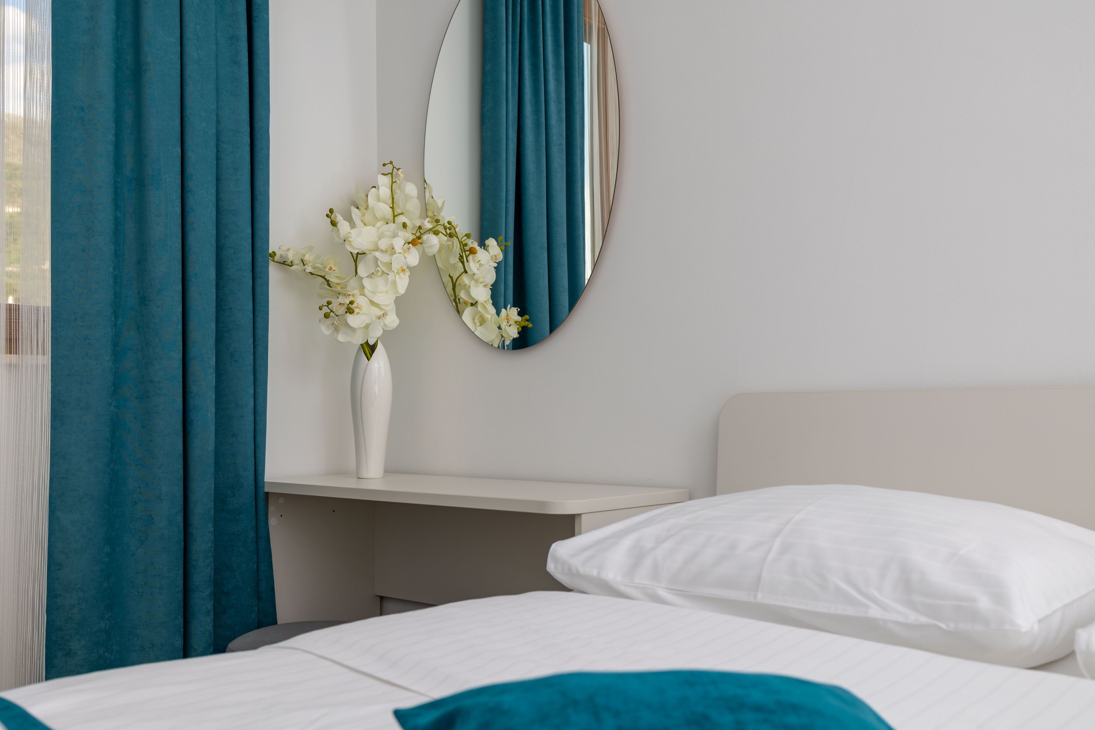 Une chambre d’hôtel avec un lit, des oreillers blancs, un vase blanc avec des fleurs, un miroir et des rideaux de couleur turquoise.