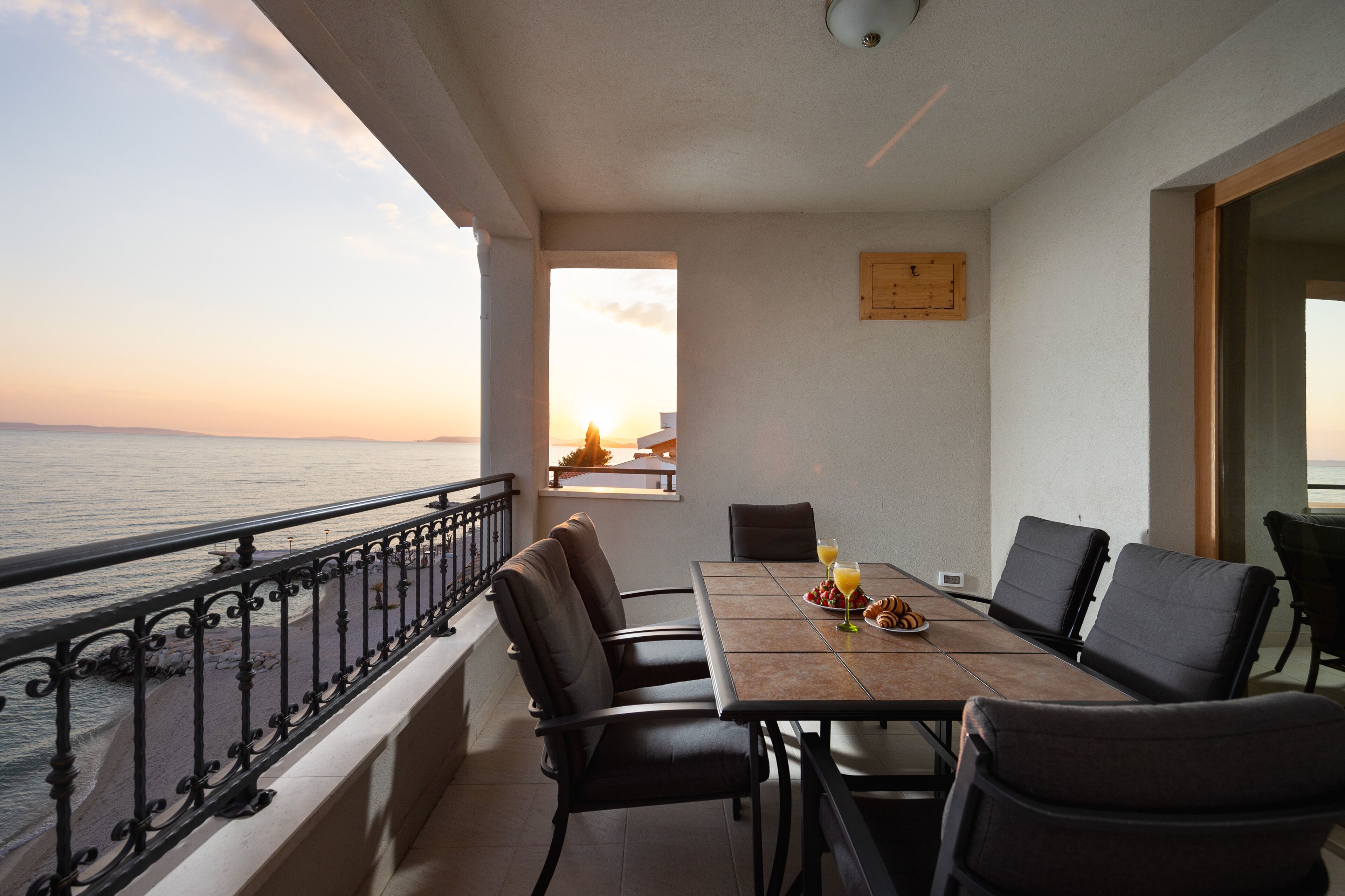 A balcony with a view of the sea, a table with chairs, and breakfast items.