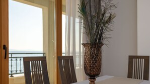Une table à manger ornée d’un vase contenant des plantes séchées, des chaises en bois et une vue sur la mer, visible à travers une porte vitrée.