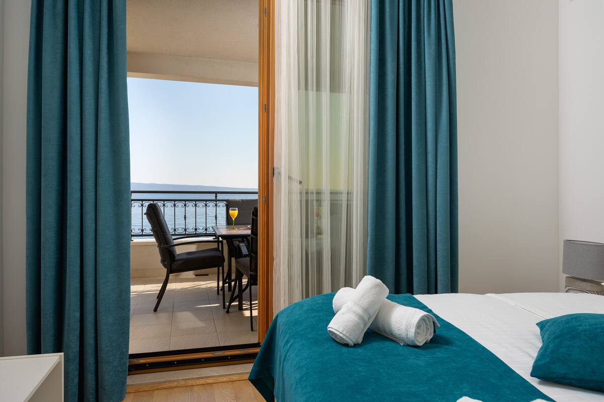 Ett hotellrum med en balkong, en säng med turkosfärgade sängkläder och utsikt över havet.