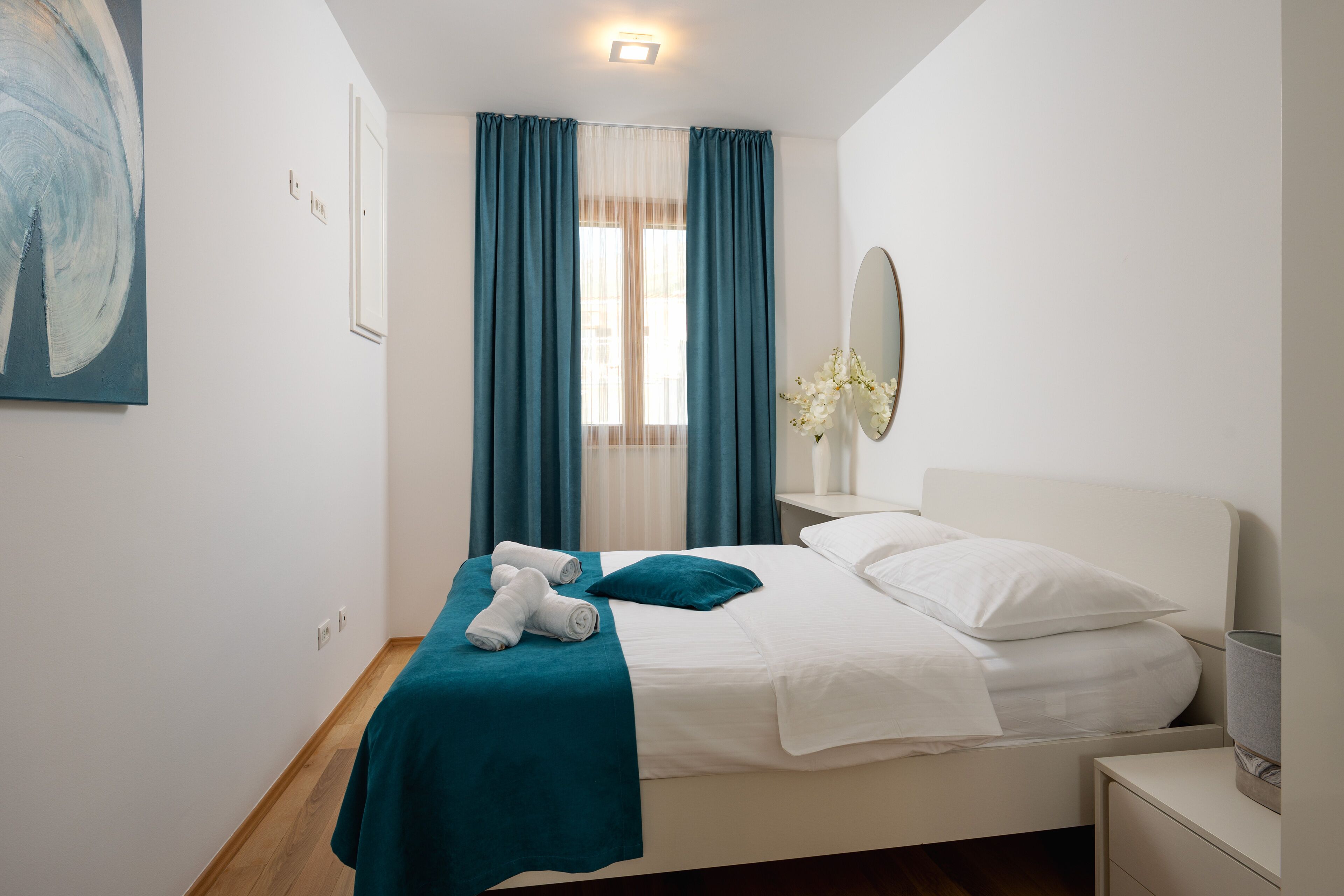 Un lit bien fait, avec du linge de lit blanc et un plaid turquoise, une table de chevet avec une lampe, et une grande fenêtre avec des rideaux bleus.