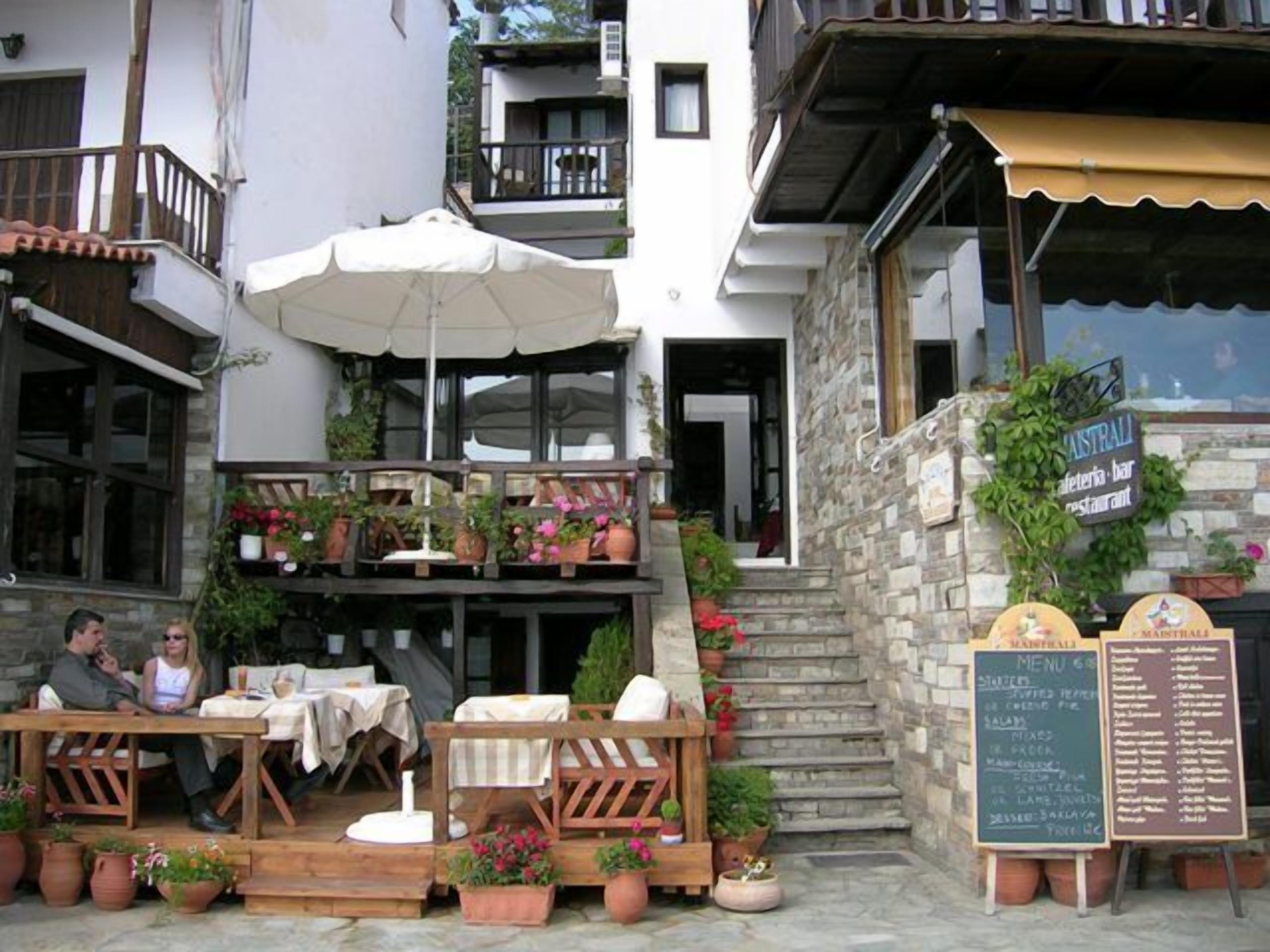 An outdoor seating area with wooden tables and chairs, surrounded by potted plants and a large umbrella.