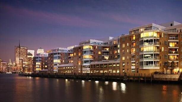 A waterfront hotel with multiple floors, balconies, and a view of the city skyline at dusk.