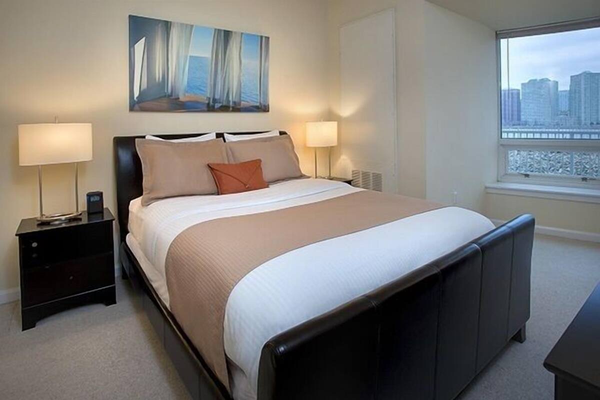 A neatly arranged bedroom with a bed, two bedside tables, a painting, and a window with a city view.