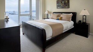 A bedroom with a large bed, a desk, and a view of a cityscape through large windows.