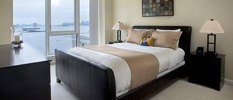 A bedroom with a large bed, a desk, and a view of a cityscape through large windows.
