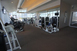 Een sportschool met diverse fitnessapparaten, grote ramen en een tapijtvloer.