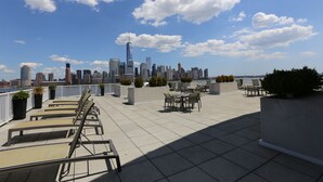 A rooftop terrace with lounge chairs and tables, offering a view of a city skyline and a body of water.