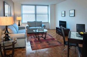 A modern living room with a sofa, a coffee table, a TV, and a dining area. - Premier Furnished Apt at Grove Pointe (Jersey City)