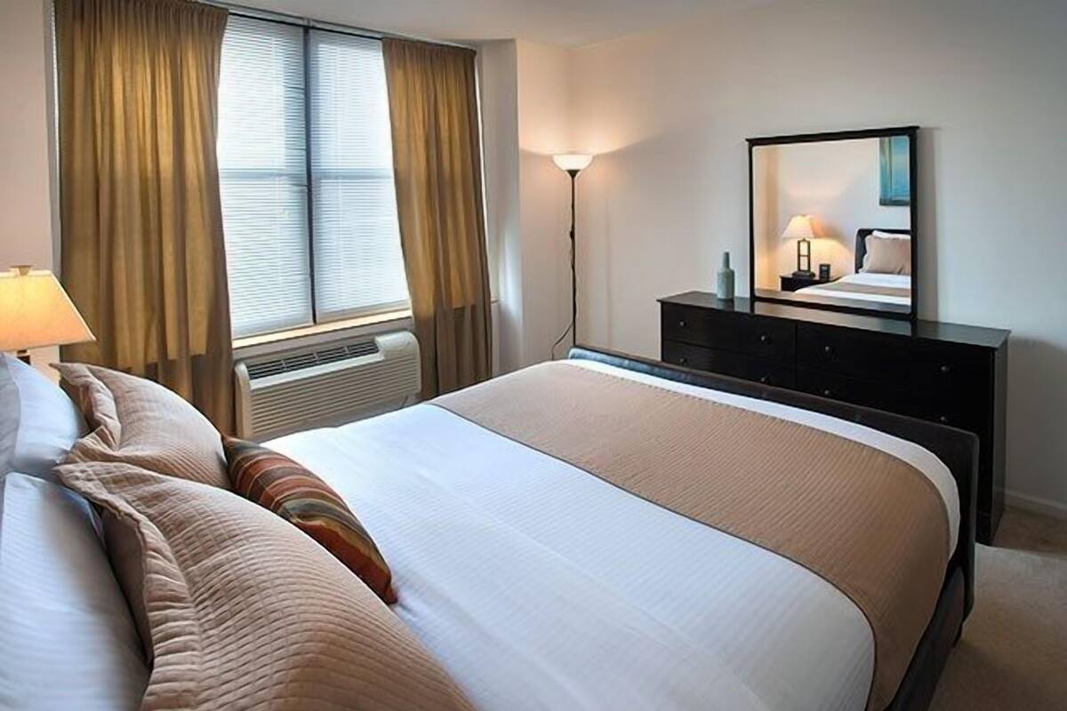 Habitación de hotel ordenada con una cama grande, un aparador, un espejo y una ventana con cortinas.