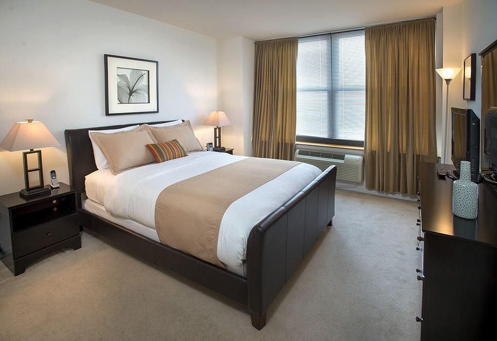 Una habitación de hotel con una cama grande, dos lámparas de noche, un cuadro enmarcado en la pared, una ventana con cortinas y un escritorio con lámpara.