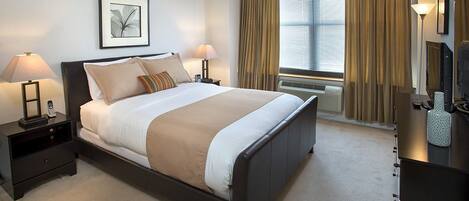 Una habitación de hotel con una cama grande, dos lámparas de noche, un cuadro enmarcado en la pared, una ventana con cortinas y un escritorio con lámpara.