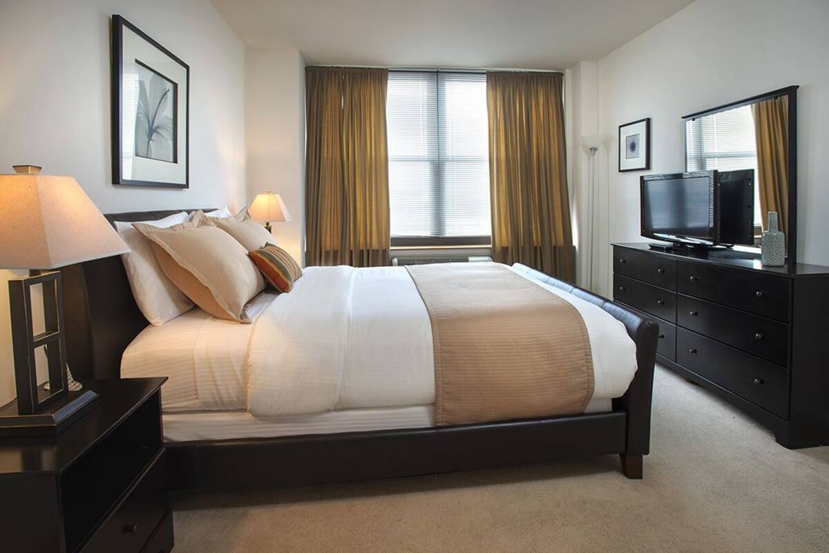 Habitación de hotel ordenada con una cama grande, lámparas de noche, un televisor de pantalla plana, un aparador y una ventana con cortinas.