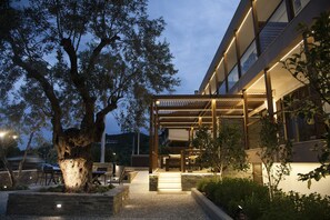 Un bâtiment moderne doté d’un espace extérieur couvert avec des sièges, d’un grand arbre et d’un chemin en gravier.