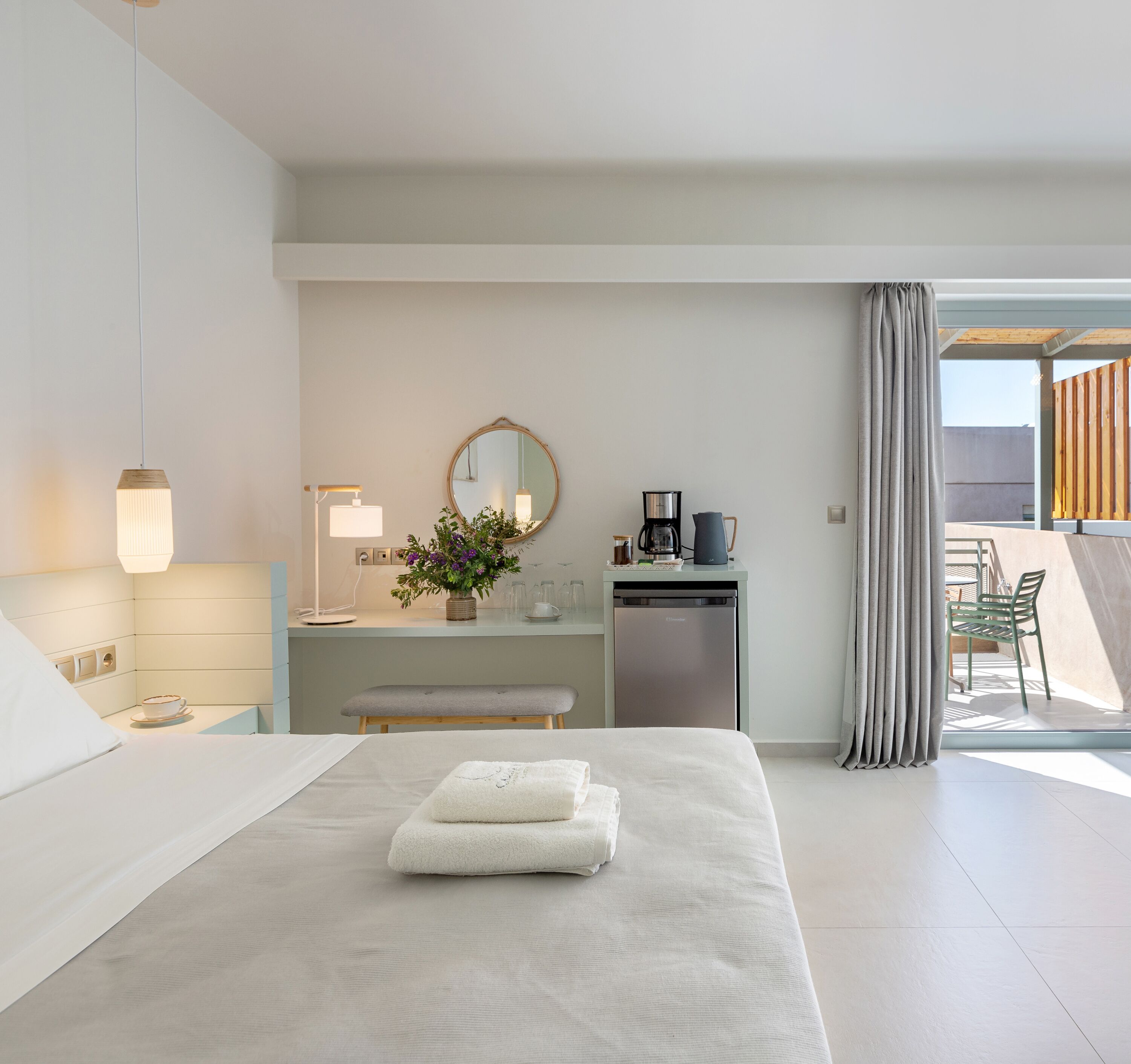 A modern bedroom with a large bed, bedside table, lamp, mirror, and a small kitchenette area with a coffee maker and kettle.