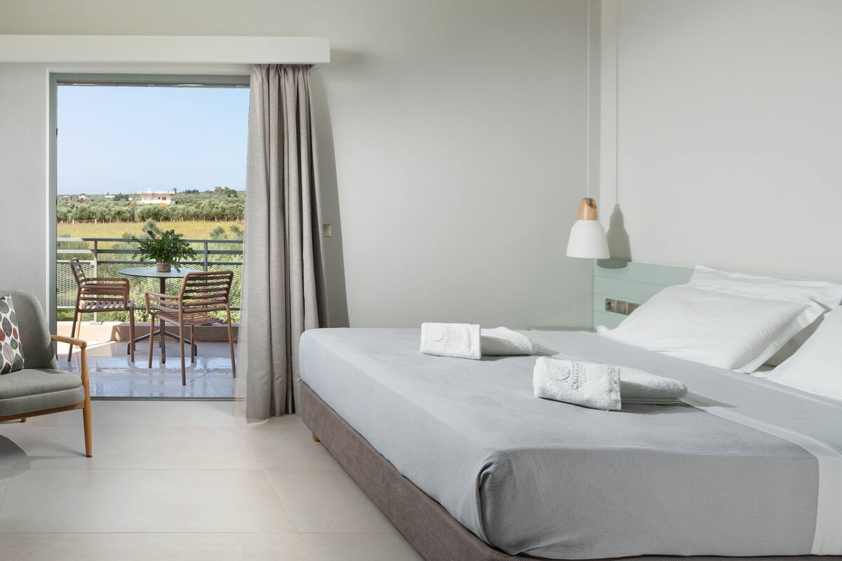 A modern hotel room with a large bed, a balcony with outdoor furniture, and a view of the countryside.
