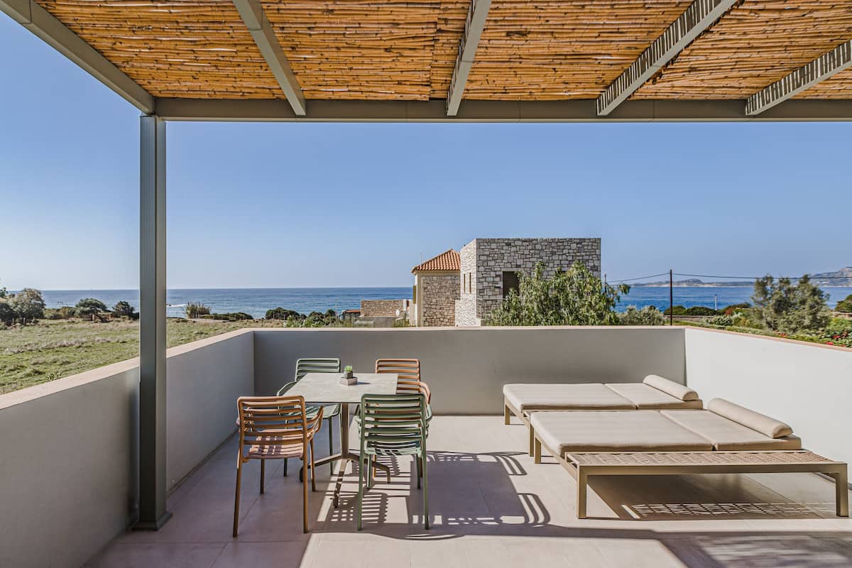 A covered outdoor seating area with a table and chairs, a sofa, and a view of the sea and a stone building in the background.