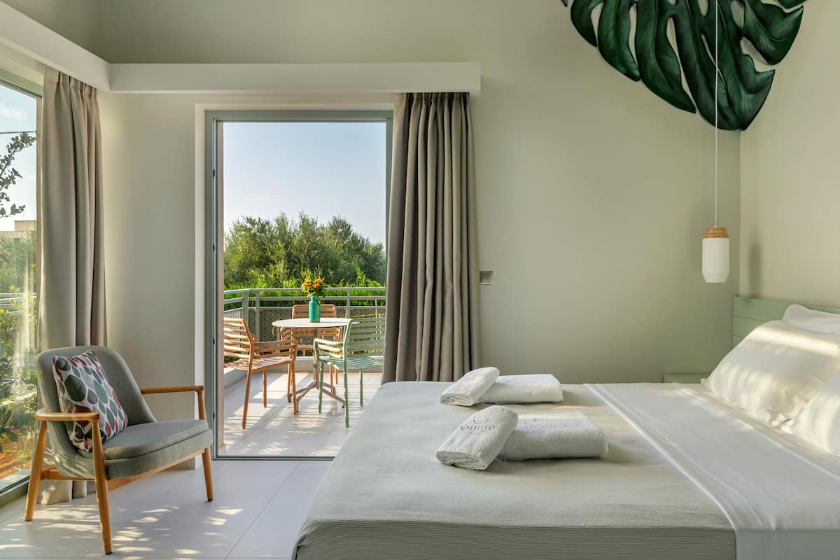 A modern hotel room with a large bed, a balcony with a table and chairs, and a view of greenery.
