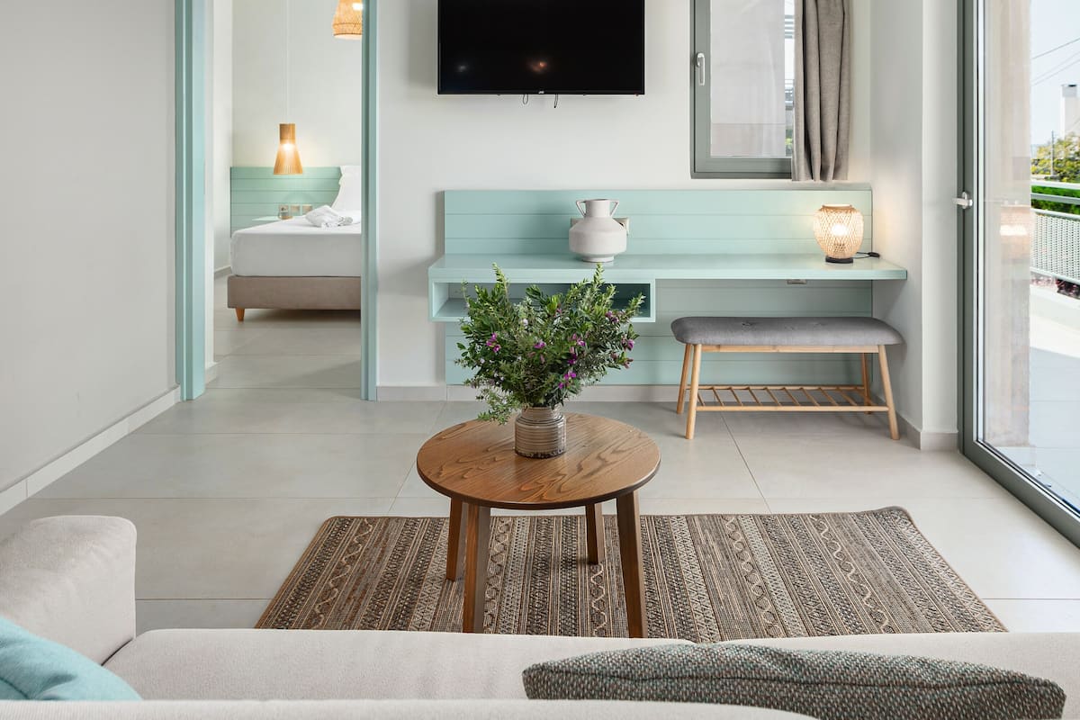A modern hotel room with a sofa, a small table with a plant, a bench, a TV, and a large window.