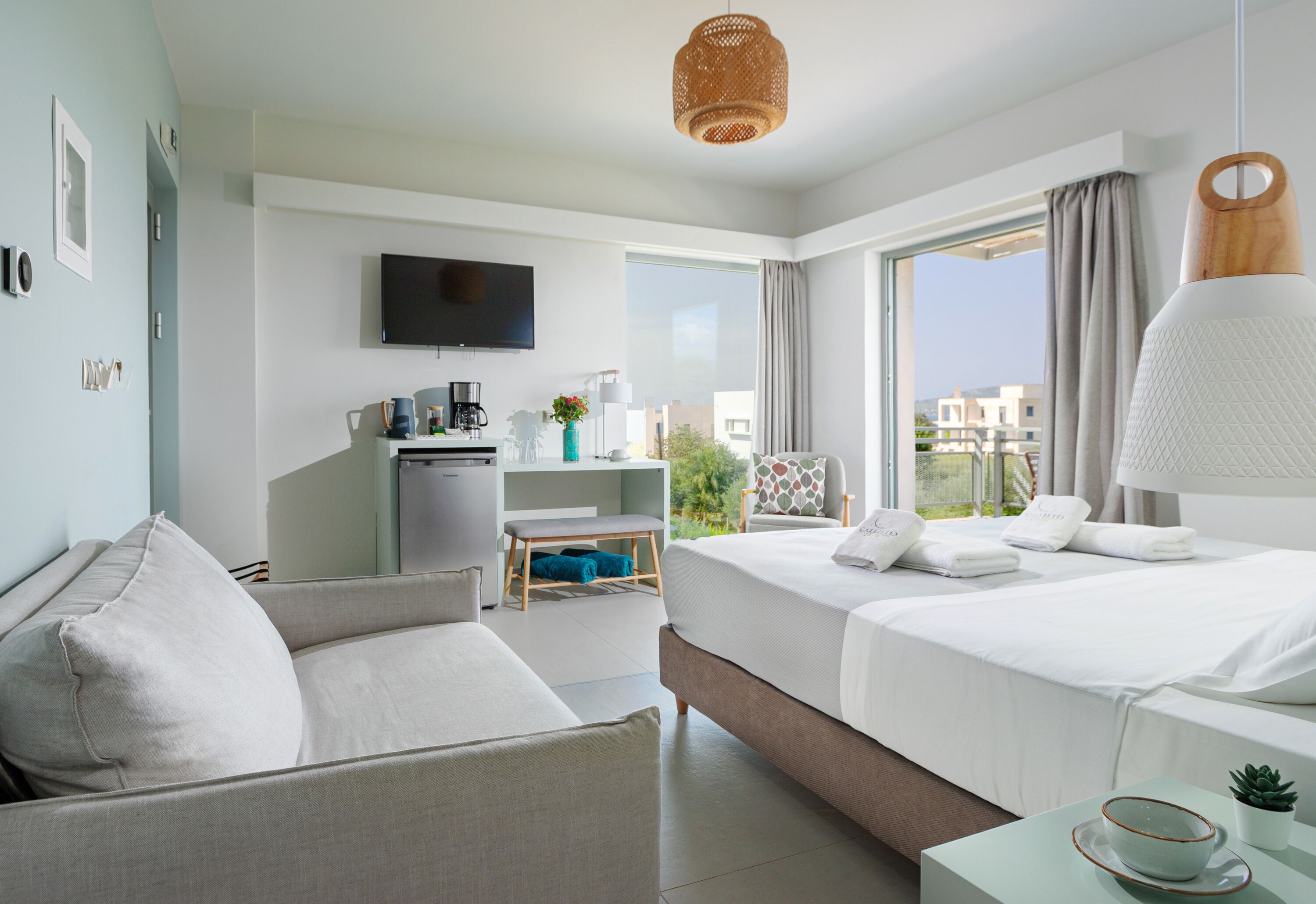 Una habitación de hotel moderna con una cama, un sofá, un escritorio con una computadora, un televisor y un amplio ventanal con vistas a edificios.