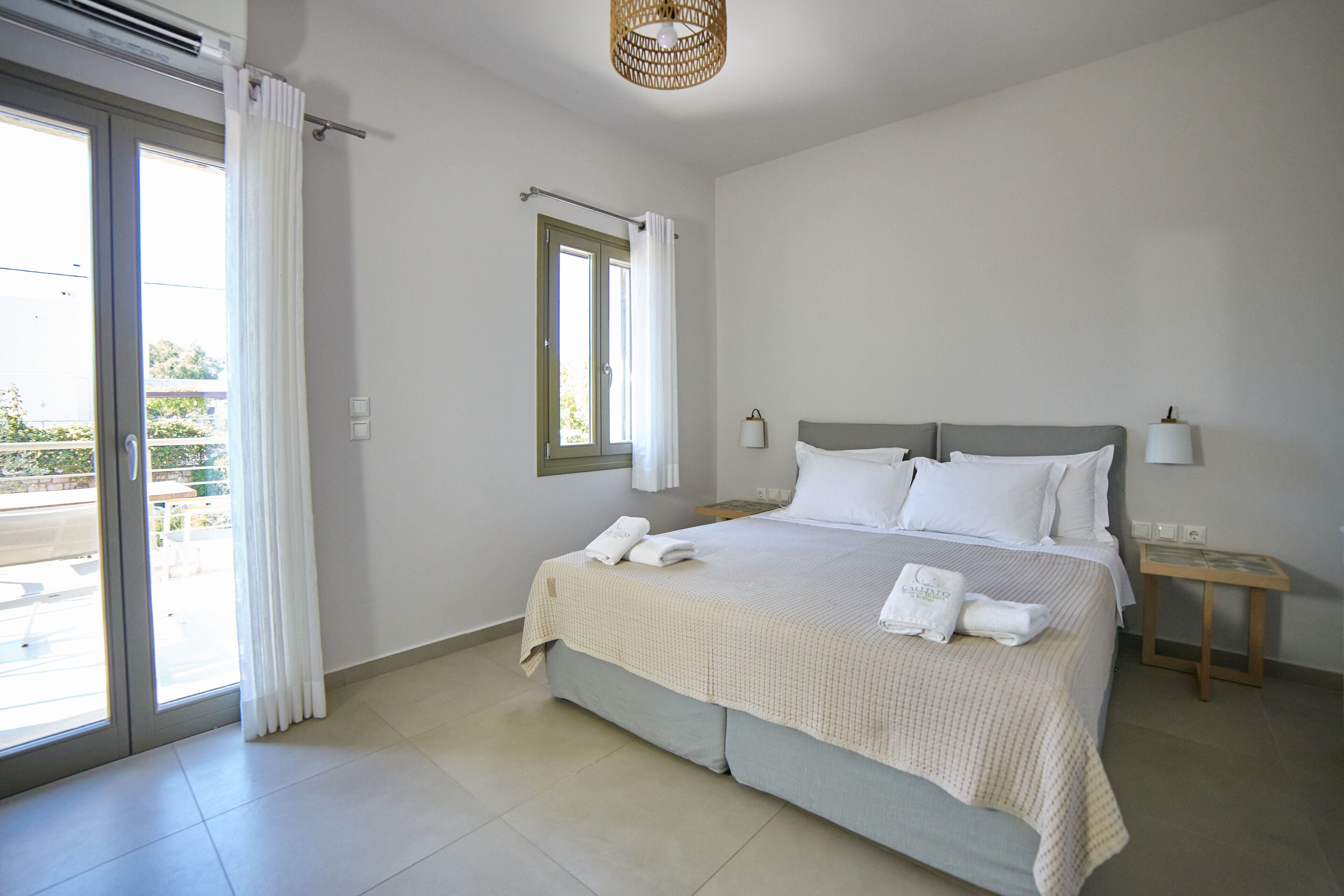 A bedroom with a bed, a nightstand, a window with curtains, and a view of the outdoors.