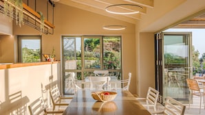 Un comedor moderno con una mesa larga, sillas blancas y un banco de madera. Hay ventanas grandes y vistas a zonas verdes.