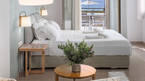 Un dormitorio con una cama grande, una mesita de noche, una lámpara, una pequeña mesa con una planta y vistas al mar a través de una puerta de balcón.