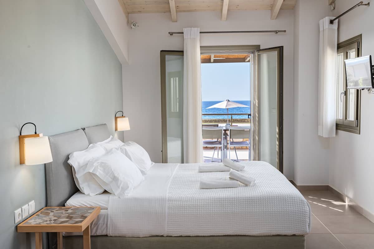 A hotel room with a bed, two bedside lamps, a small table with a chessboard, and a view of the sea through an open door.