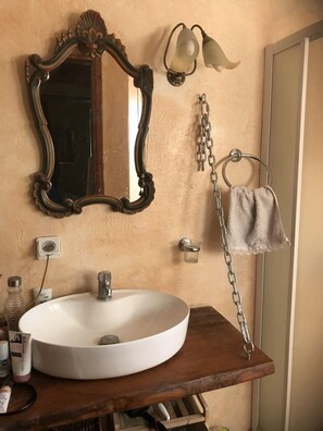 Une salle de bain avec un lavabo rond blanc, un meuble-lavabo en bois, un miroir décoratif, des appliques murales et un porte-serviettes.