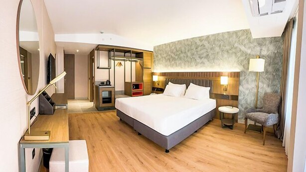 Habitación de hotel con una cama grande, un escritorio con silla, un espejo, un armario y una mesita de noche con lámpara.
