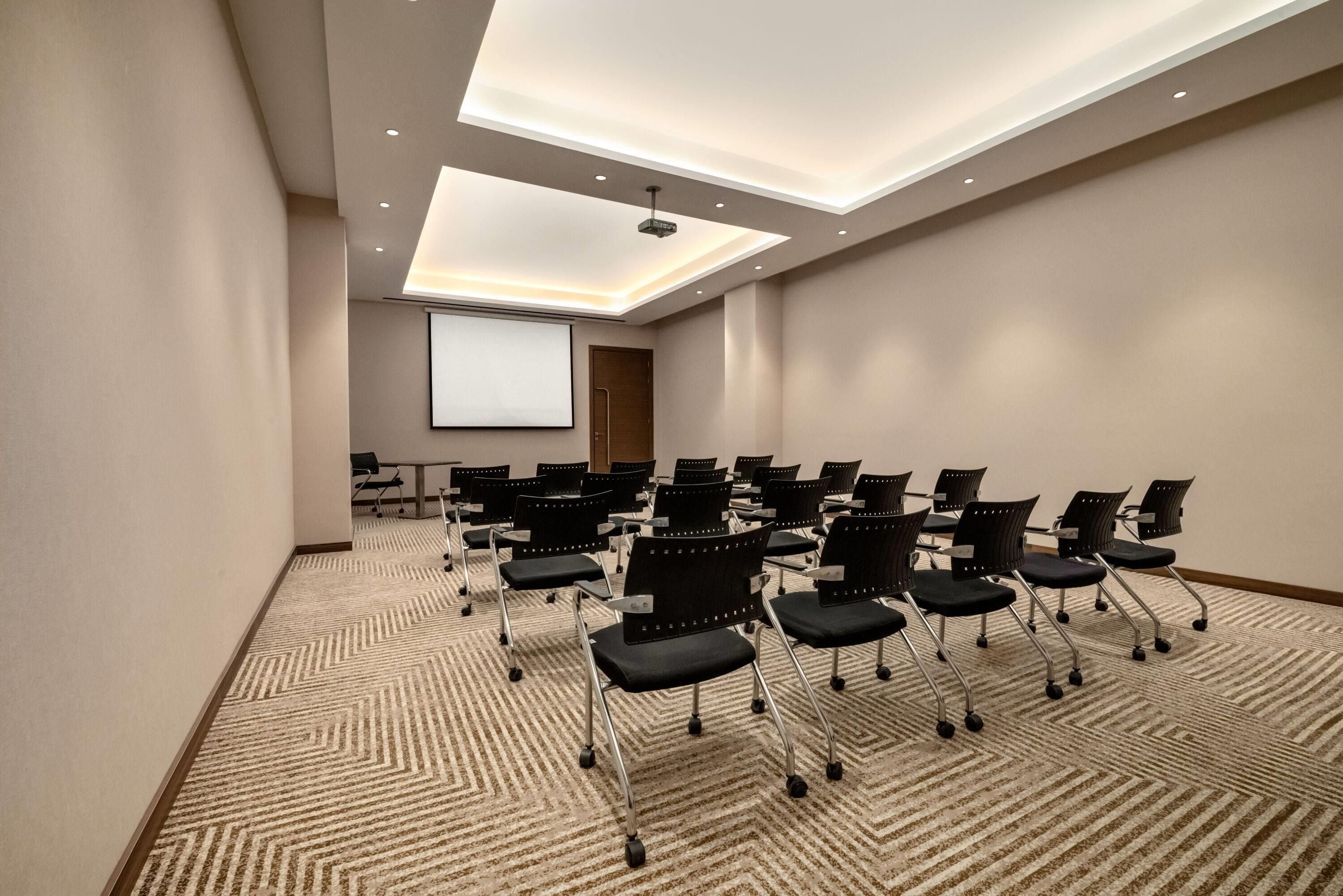 Une salle de conférence dotée d’un grand écran, de plusieurs rangées de chaises et d’un sol recouvert de moquette.