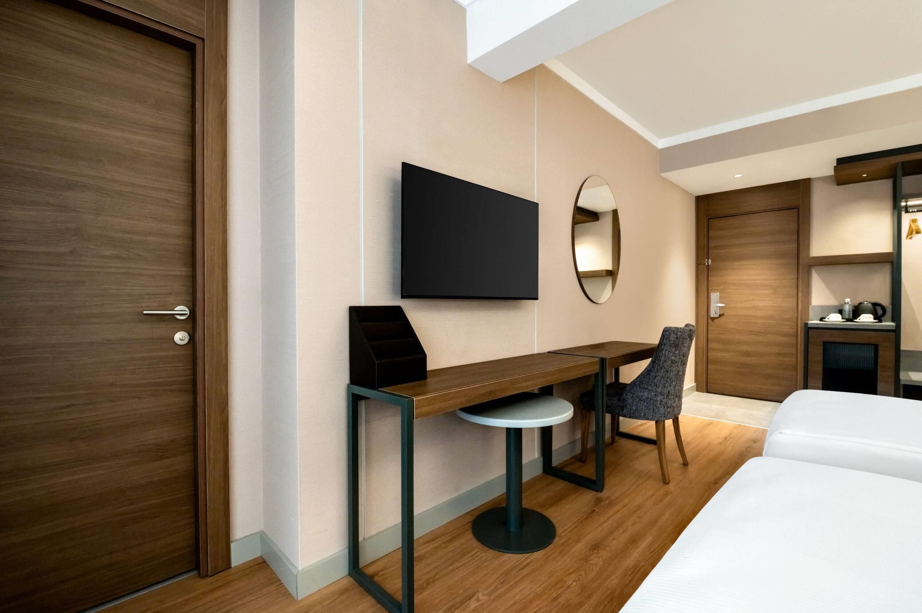 A hotel room with a wooden door, a desk with a chair, a television, a round mirror, and a bed.