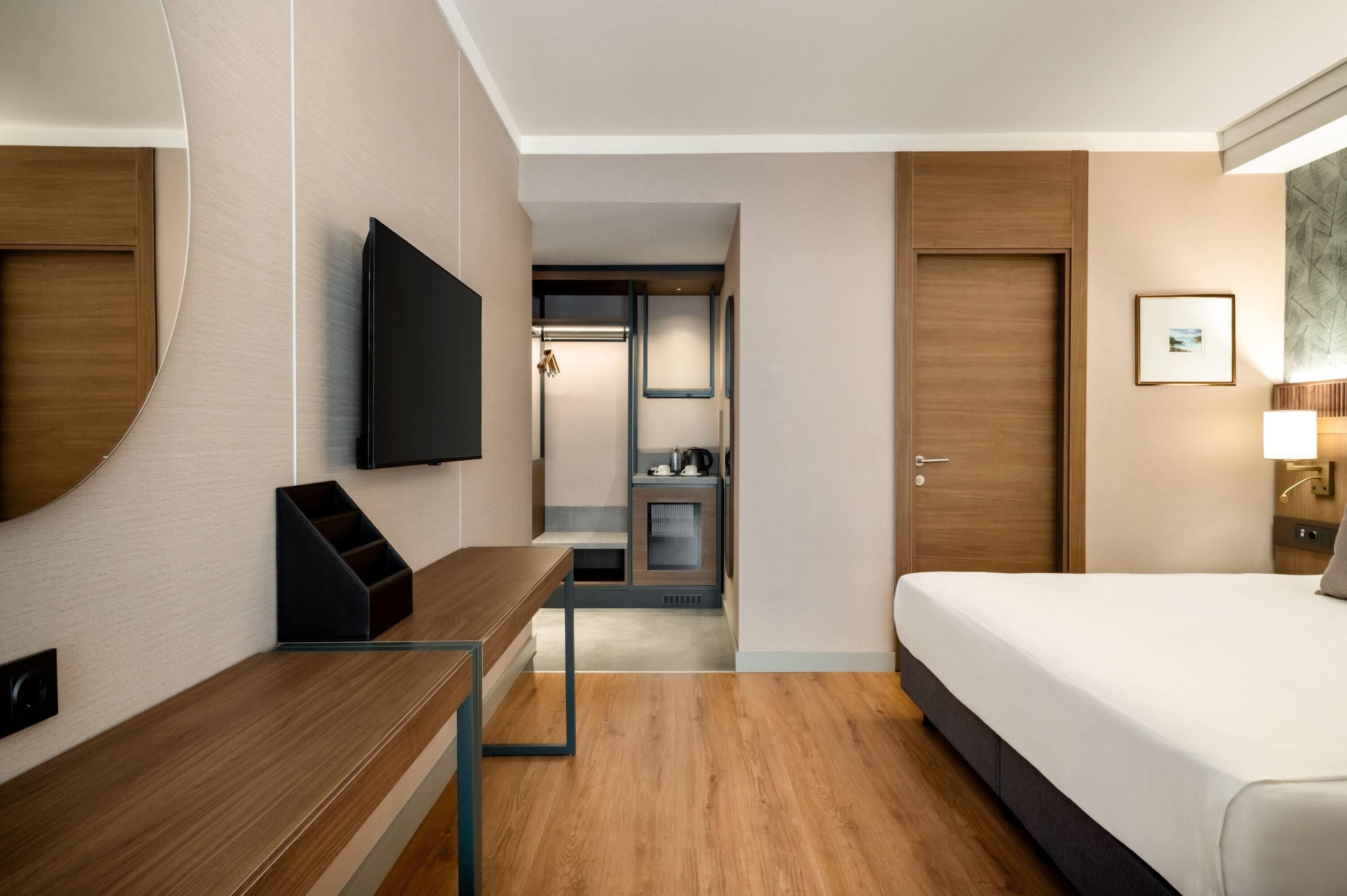 A hotel room with a bed, a desk, a television, a door, and a small kitchenette.
