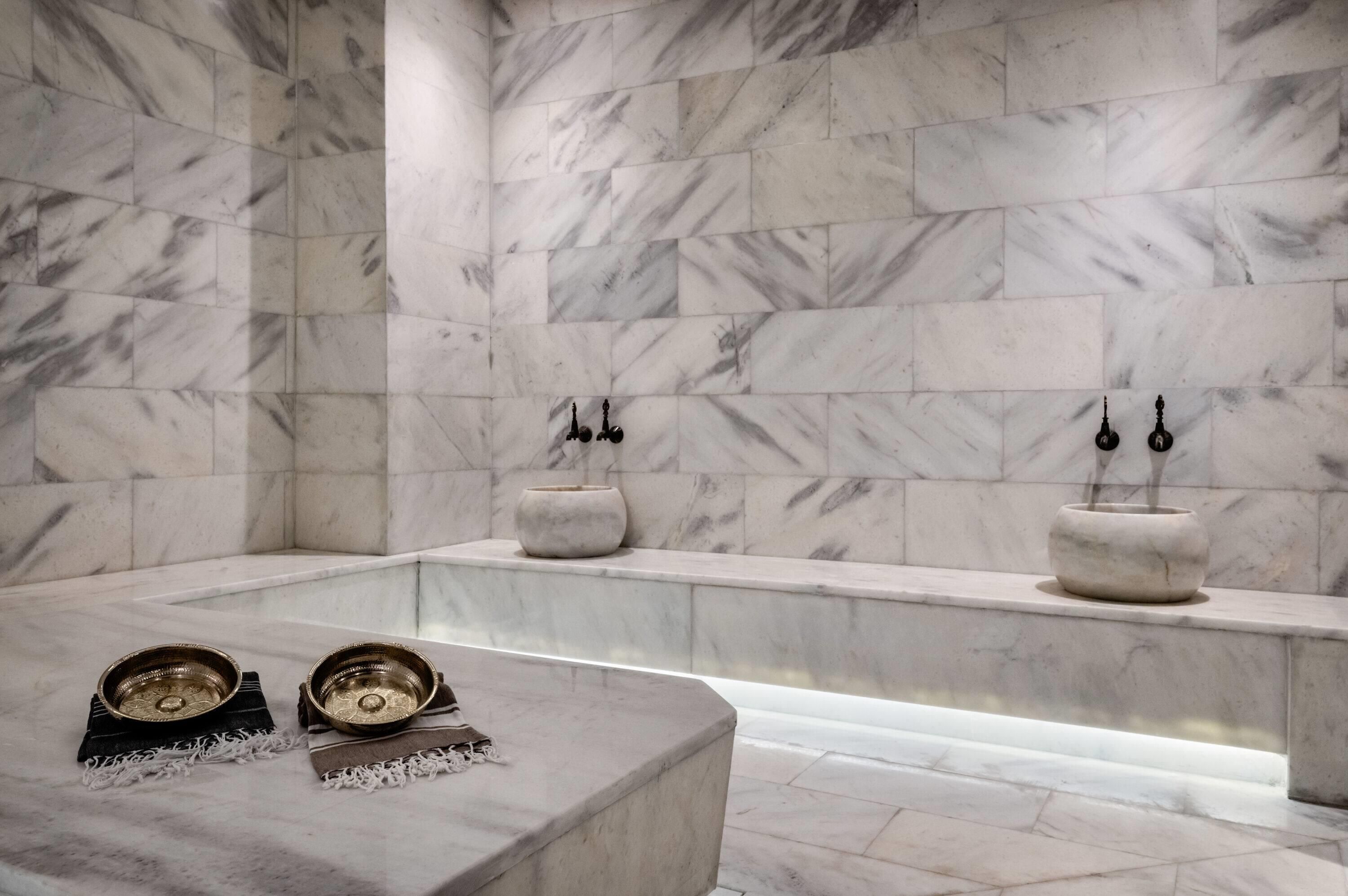 A marble bathroom with a freestanding bathtub, two wall-mounted basins, and a pair of decorative bowls on a bench.